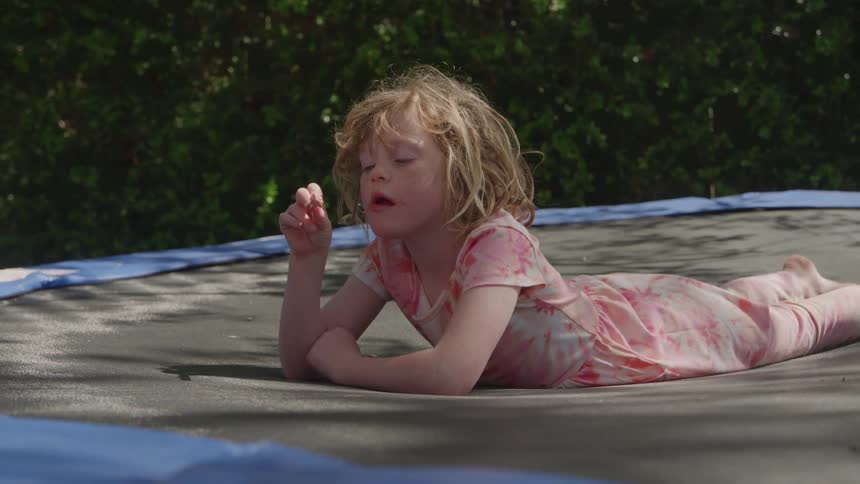 A little girl with long blonde hair lies on her stomach on a trampoline licking a red popsicle on a sunny day outdoors