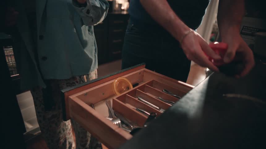 A couple works sidebyside in a modern kitchen as the woman loads or unloads a dishwasher while wearing headphones and the man puts silverware into a divided drawer stopping to smile at her
