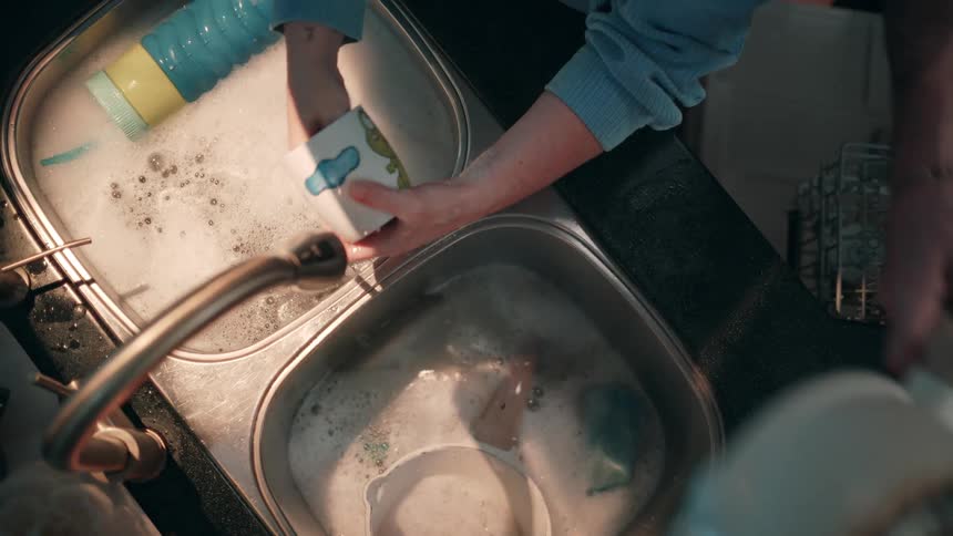 A top down view of a person wearing a blue shirt cleaning dishes and a pot in a double kitchen sink filled with soapy water
