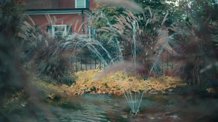 A blurred view of a water fountain with decorative grasses and yellow plants showing a woman pushing a man on a wheelchair passing a brick building in the background