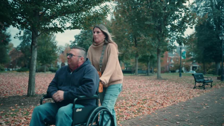 A woman pushes a man in a wheelchair along a leaf covered brick pathway in a lush public park on an autumn day
