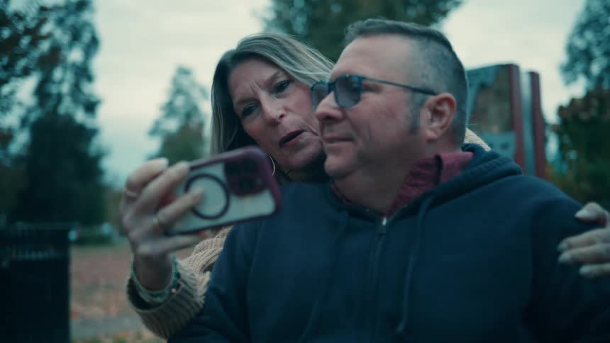 A middleaged couple is shown outdoors taking a selfie together using a smartphonethe woman has her arm around the man who is wearing glasses and they smile at the camera after sharing a quick kiss