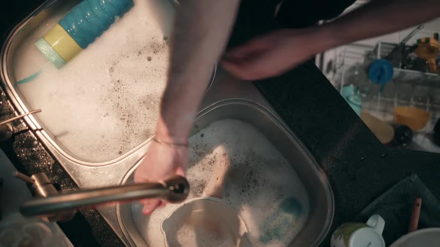 From an overhead perspective a persons arms and hands are shown actively washing darkcolored dishes in a doublebasin kitchen sink filled with soapy water using a sponge and rinsing the items under running water from a brasscolored faucet