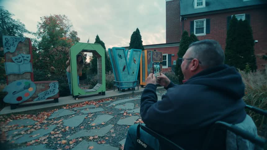 A man in a wheelchair takes a photo of a woman posing with large colorful letter shaped art sculptures on a stone pathway outside a brick building