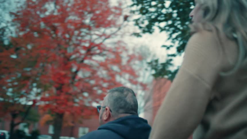 Woman pushes man in wheelchair through autumn park with colorful trees and benches in background