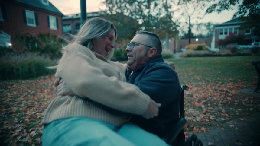 A woman sits on the lap of a man in a wheelchair as they embrace and share a passionate kiss outdoors in a park setting during the late afternoon