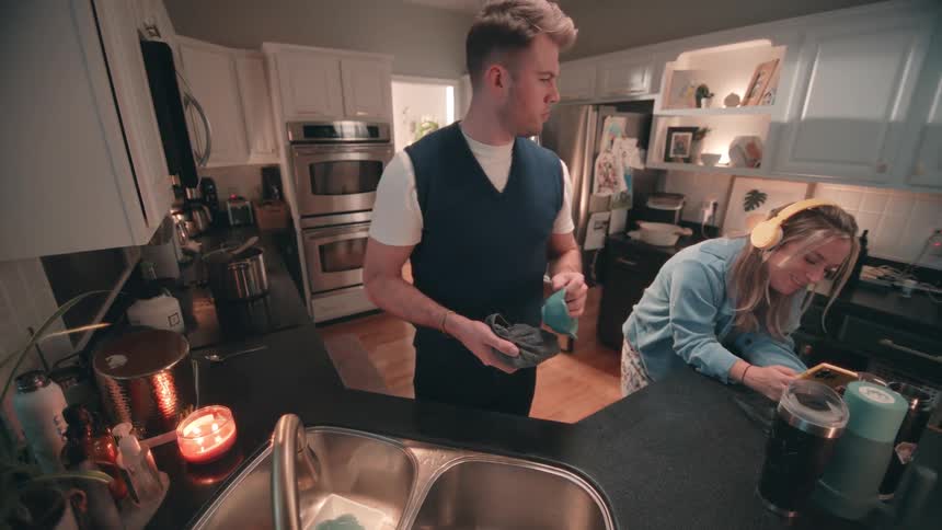 The woman listens to music while the man does dishes