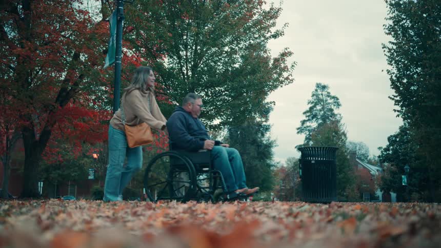 A calm woman pushes a relaxed older man in a wheelchair on a path covered in colorful autumn leaves