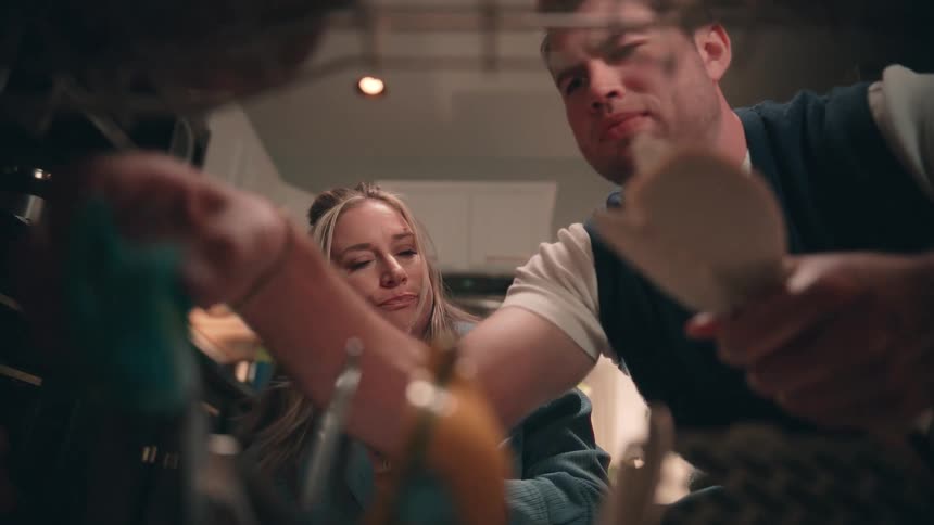 The video shot from inside a low cabinet shows a couple reaching into the space for kitchen items suggesting they are stocking or searching for supplies the woman hands the man a spoon and a plate before standing up while the man continues to rummage