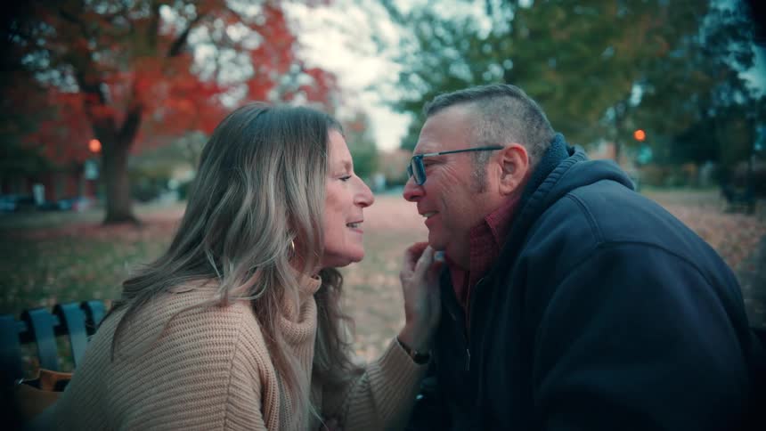 A middleaged couple sits on a public bench in the park sharing a kiss while wearing jackets surrounded by trees