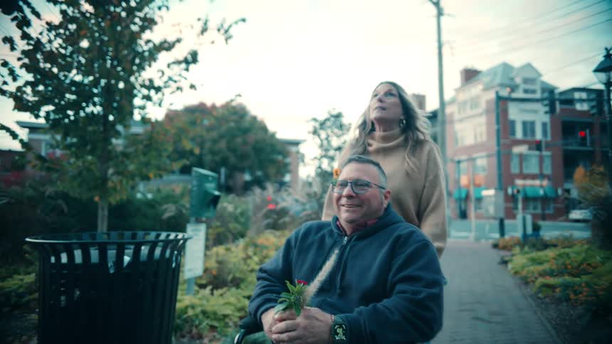 Man in a wheelchair being pushed by a woman while they walk through a treelined urban area