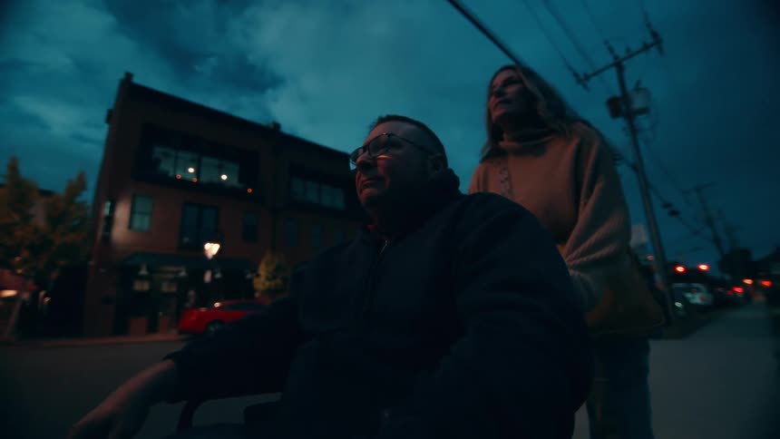 Woman pushing a man on a wheelchair on a city sidewalk with cars passing in the background