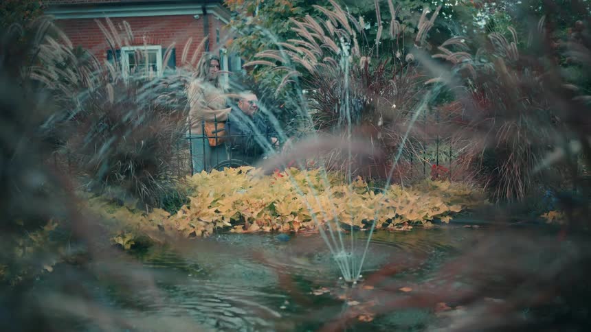 A middleaged couple spends midday in the park with the man seated in a wheelchair and the woman standing behind him as they watch a water fountain