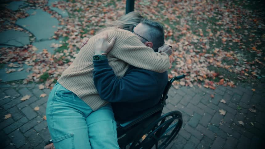 A couple is in the middle of the street at midday the woman sits on the lap of the middleaged man in a wheelchair hugging him warmly