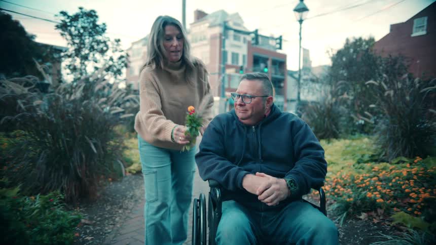A woman walks up behind a man sitting in a wheelchair on a brick sidewalk lined with plants and she gives him a small orange flower that she picked before leaning down to kiss him on the cheek resulting in both people smiling