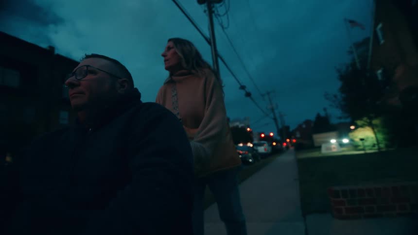 A woman pushes her husband who is seated in a wheelchair along the sidewalk in the evening