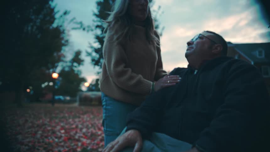 A woman in a beige sweater and jeans stands next to a man seated in a wheelchair outdoors leans down to talk to him briefly kisses him and touches his chest affectionately in a park setting during twilight
