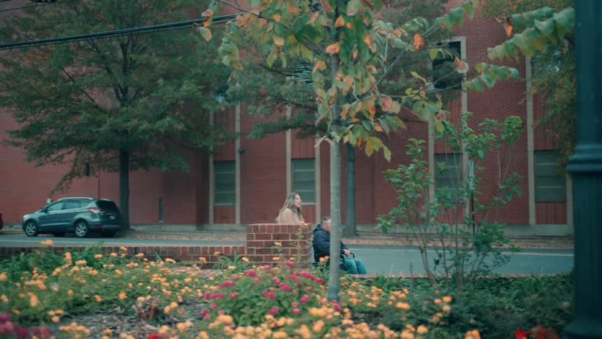 A woman is seen helping push a man in a wheelchair along an outdoor path next to a brick buildingthe foreground features a bright flowerbed with yellow and red blossoms framed by trees suggesting an autumn season