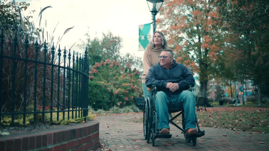 A woman in a tan sweater pushes a man seated in a black wheelchair along a paved walkway through a public park passing a low brick wall and a wrought iron fence in the foreground with autumn trees and a lamppost visible