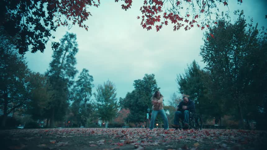 A wide lowangle shot in an autumn park shows a woman standing next to a man seated in a wheelchair first tossing a handful of fallen leaves into the air before bending down to embrace him warmly