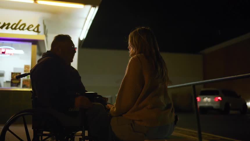 Man with glasses and sitting on a wheelchair kissing a woman sitting down in front of him next to an ice cream shop in an urban area outdoors during night time