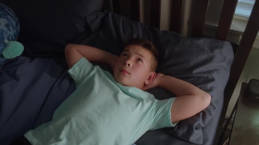 Contemplating young boy laying in bed with his arms behind his head
