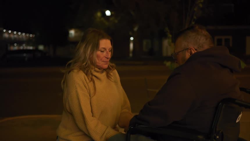 A laughing woman in a beige sweater is talking to a man in a wheelchair wearing a dark hooded sweatshirt and glasses outside at night