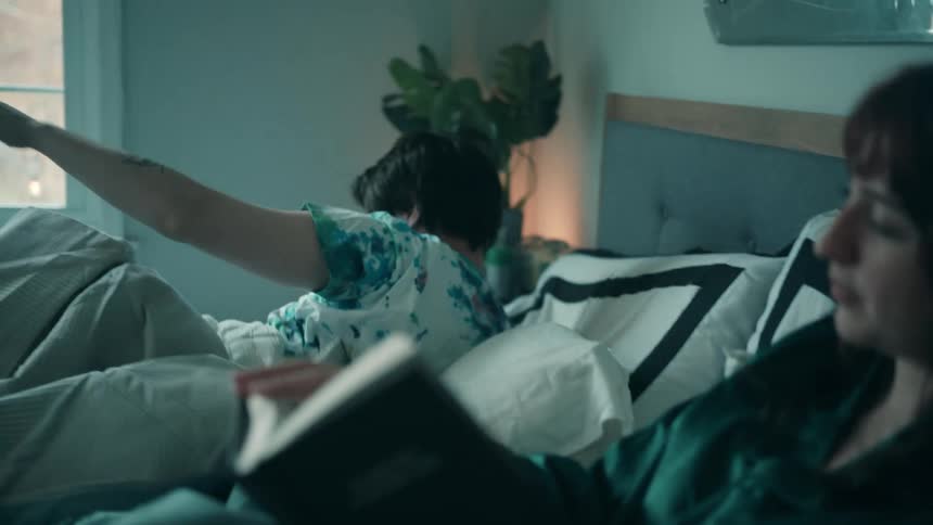 A woman with dark hair sits up in bed reading a book in the foreground while a man sleeping beside her suddenly throws off the covers and sits up stretching in the brightly lit bedroom that features a large window