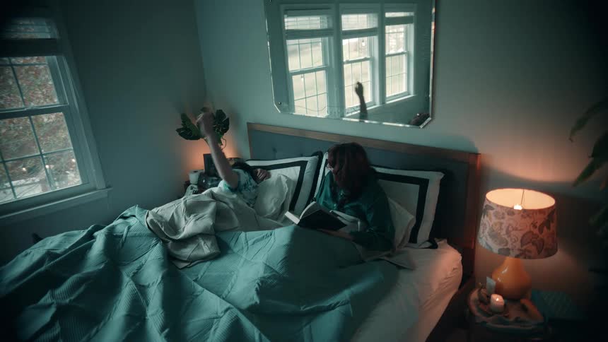 Person reads in bed beside someone sleeping under soft bedroom lighting