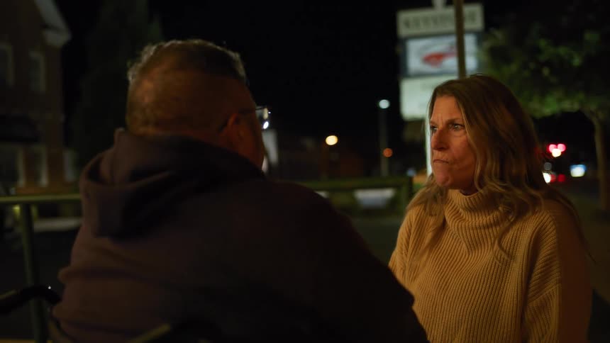 A woman in a yellow turtleneck sweater earnestly talking to a man whose back is mostly to the camera as they have a focused conversation outdoors with streetlights and businesses dimly lit behind them