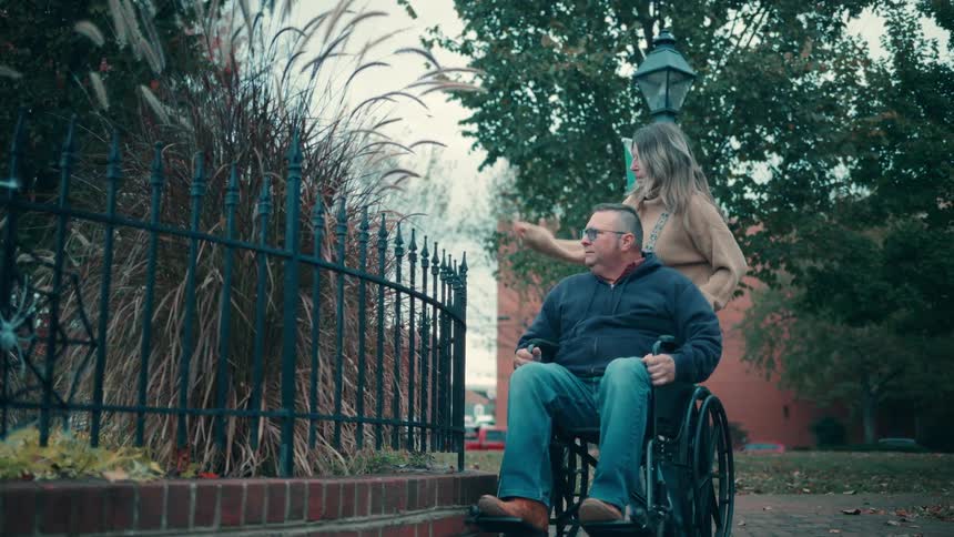 Person in a wheelchair is accompanied by another while observing vegetation in an urban park