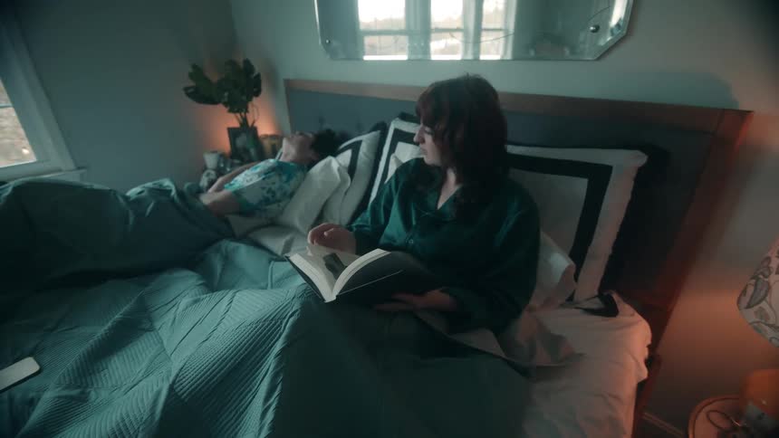 A woman sits up reading a book in bed while a man next to her finishes looking at his phone and goes to sleep in a dimly lit bedroom