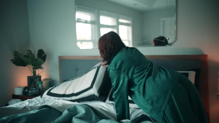 The video shows a person with reddishbrown hair wearing a green garment making a bed in a dimly lit bedroom they are arranging pillows and smoothing out a tealcolored blanket or duvet with light from a window reflecting in a mirror above the headboa
