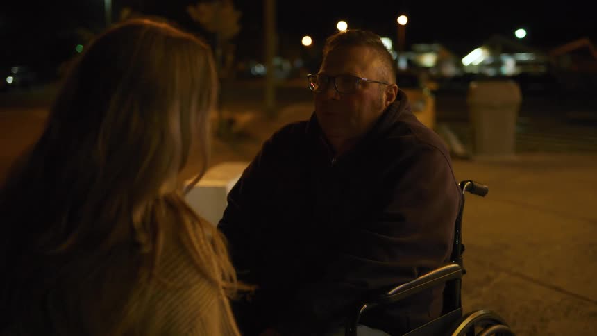 A woman with long lightcolored hair is speaking to a man who is sitting in a wheelchair the scene takes place outdoors at night with streetlights visible in the background creating an intimate atmosphere