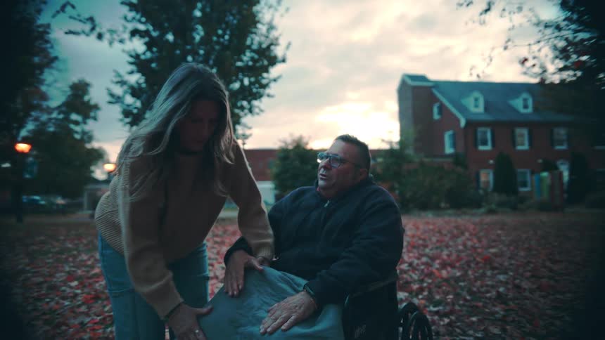 A middleaged couple is outdoors at midday the man sits in a wheelchair while the woman covers his feet with a blanket kisses him and prepares to roll him across the street