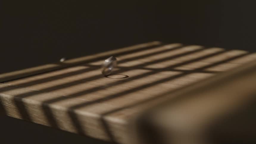 A metal ring is shown spinning and wobbling on a grooved wooden cutting board striped shadows possibly from blinds fall across the board creating a strong contrast of light and dark