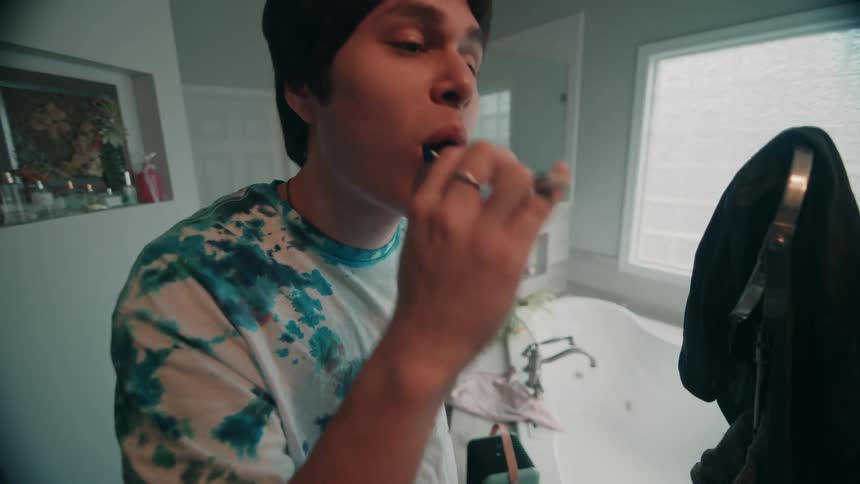 A close view of a young man with dark hair and a tie dye shirt brushing his teeth in a brightly lit modern bathroom