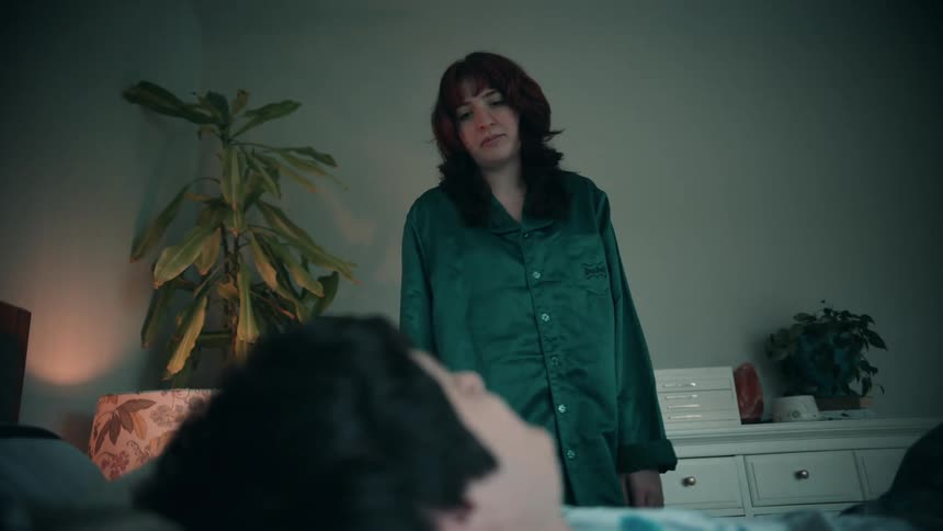 A woman in green pajamas stands by a bed looking down at a person lying beneath the covers with the shot taken from the perspective of the person in bed in a dimly lit bedroom setting
