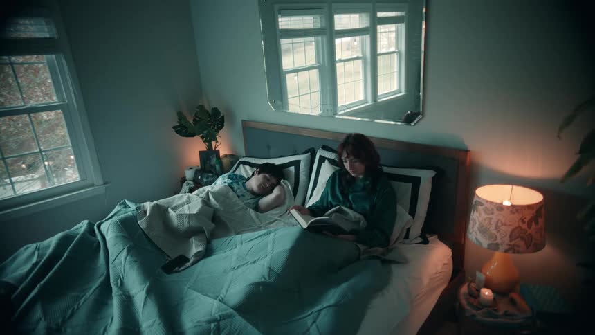 Person sleeping beside someone reading a book in bed in a cozy bedroom