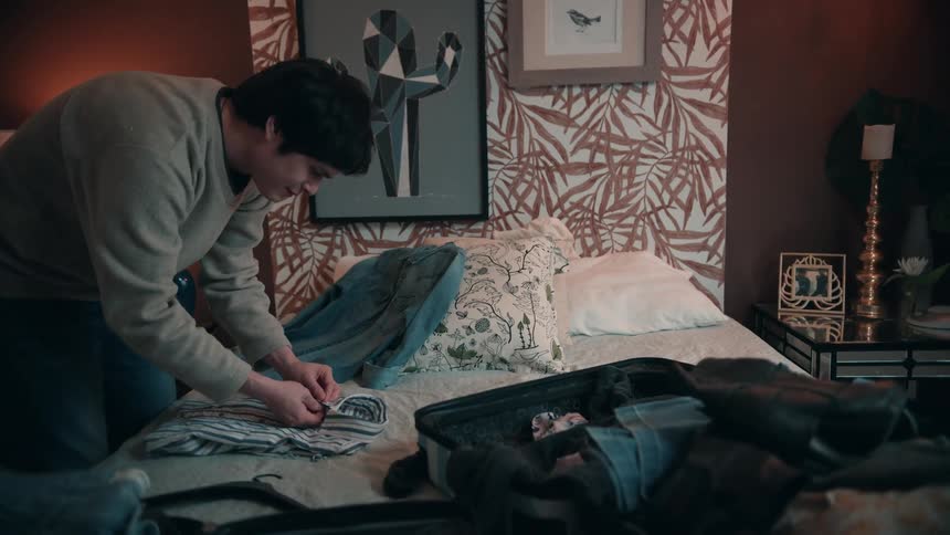 Man folds clothes on the bed while organizing items inside the open suitcase