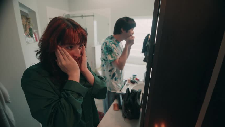 A young couple is in the bathroom getting ready the woman faces the mirror applying makeup while the man brushes his teeth