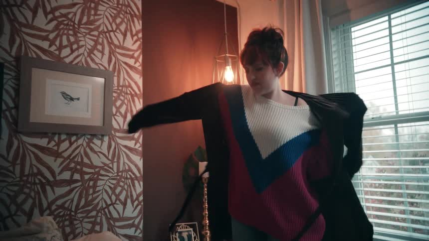 A woman gets dressed in a bedroom as a man is busy with the bed