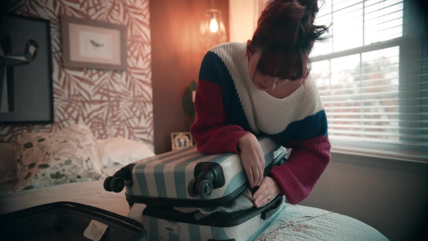 The woman leans on the luggage in order to close it