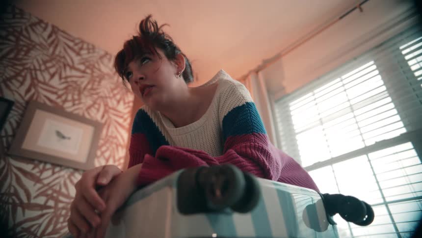 A low angle view shows a woman in a colorful sweater leaning over a suitcase struggling to close it in a brightly lit room with patterned wallpaper
