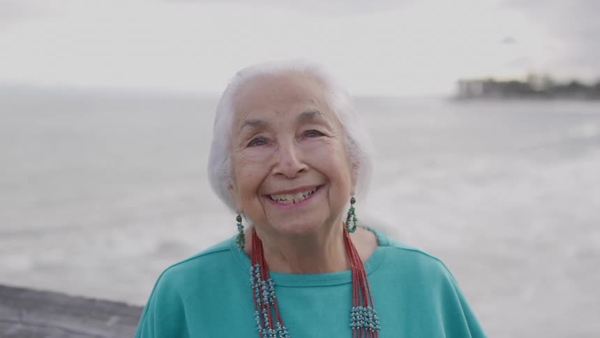 A close view of a beautiful older woman with white hair wearing beaded necklaces and earrings smiling happily in front of a misty gray ocean