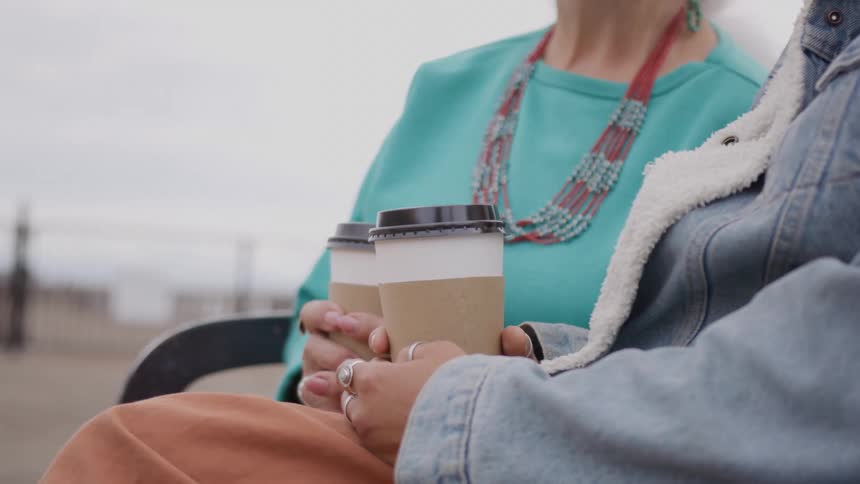 A close view of two people sitting outdoors holding takeaway coffee cups with one wearing a turquoise top and a red beaded necklace and the other a denim jacket