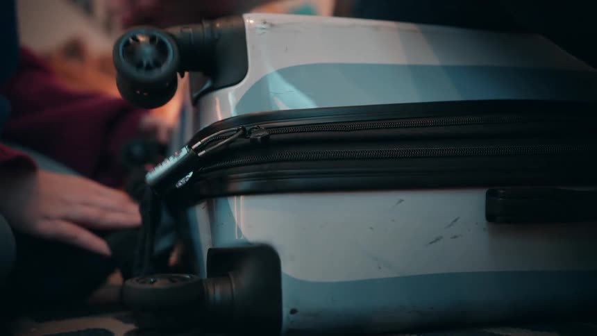A close view of a person zipping up and locking a white and blue rolling suitcase on a dark carpeted floor