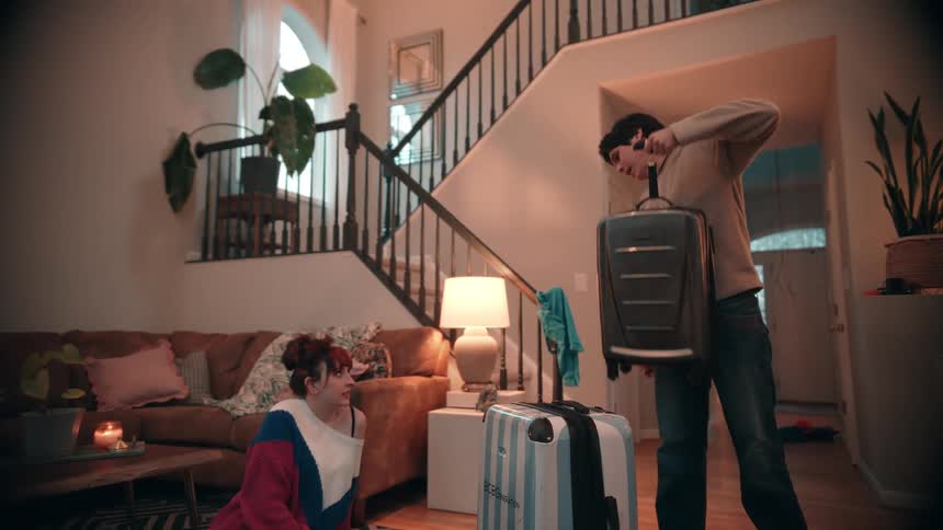 A man and a woman stand by several suitcases near a staircase in a warmly lit living room preparing to leave on a trip