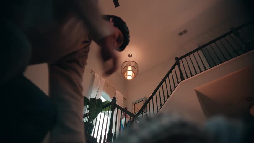 The video is shot from a lowangle perspective looking up a carpeted staircase with a black metal railing inside a housein the foreground a person is seen bending over holding and dropping or throwing down some dark clothing while another person