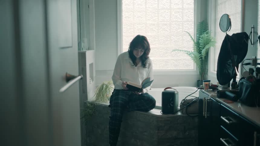 Teen girl sitting down and reading a book while a teen boy searches on drawers and then walks away to another room indoors during day time
